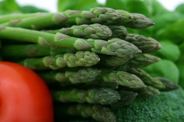 A close-up of a fresh bunch of asparagus alongside a ripe tomato and leafy greens showing vibrant colors. Perfect for promoting healthy eating, organic produce, farm-to-table concepts, and nutrition. Ideal for use in food blogs, gardening websites, recipes, marketing materials for farmers' markets and grocery stores.