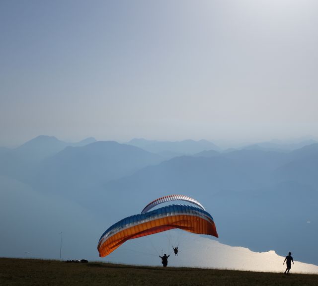 Two paragliders are seen launching off a mountain with a hazy blue horizon and misty hills in the background. Ideal for use in adventure, outdoor sports, and travel advertisements. It evokes feelings of freedom, tranquility, and the exhilaration of extreme sports. Suitable for promoting paragliding experiences, scenic travel destinations, or nature-inspired activities.