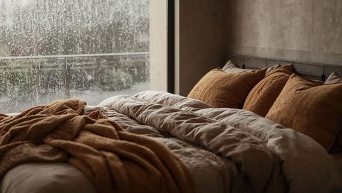 Rain falling outside cozy bedroom with unmade bed, rust pillows and textured duvet