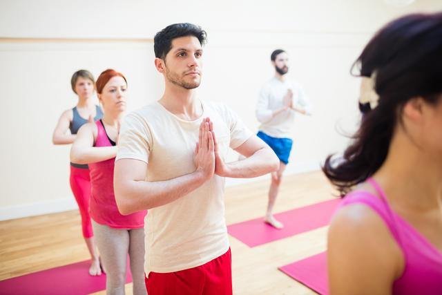 Group of individuals engaging in a tree pose exercise during a yoga class in a fitness studio. Ideal for promoting wellness, balance, and mindfulness, this image is perfect for use in fitness blogs, yoga studio advertisements, wellness programs, and health-related marketing materials.