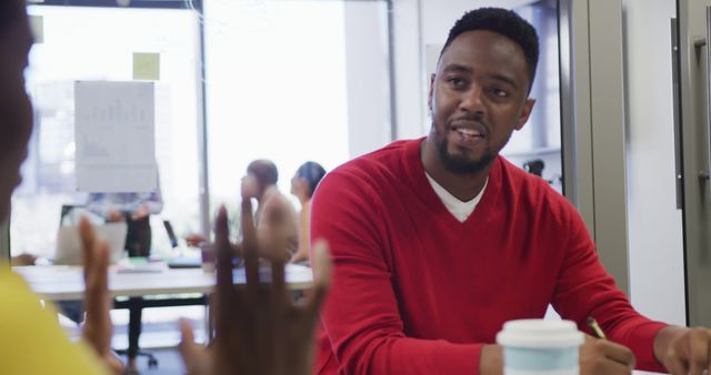 Young man in red sweater engaged in discussion with co-worker in modern office setting. Others are seen in background working collaboratively. Ideal for themes of teamwork, communication, inclusive workplace, professional environment, modern workspace.