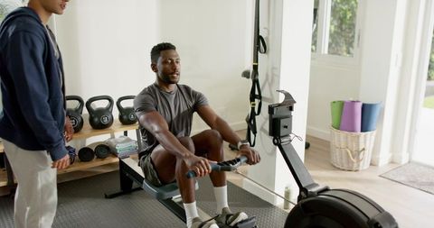 Black male client rowing on erg while trainer coaching in sunlit home gym with kettlebells
