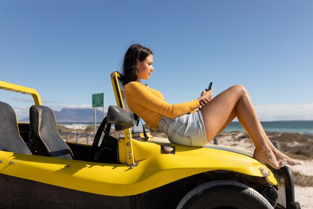 Woman lying on yellow beach buggy using smartphone on a sunny beach. Ideal for themes related to summer holidays, road trips, relaxation, technology use outdoors, and coastal adventures. Perfect for travel blogs, vacation advertisements, and lifestyle magazines.