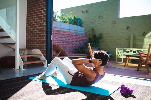 Young African American man performing crunches on an exercise mat at home. Sunlight streams through the open door, illuminating the space. Ideal for use in articles or advertisements related to home fitness routines, healthy lifestyles, and indoor exercise tips.