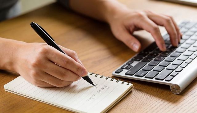 Hands writing notes while typing on sleek wireless keyboard on wooden desk, close-up