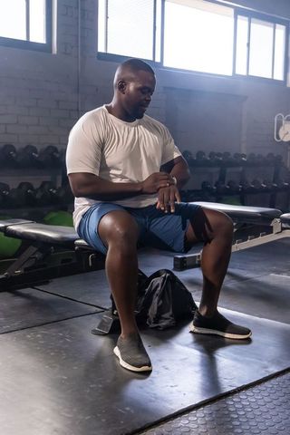 African american man sitting on bench checking smartwatch after workout in industrial gym