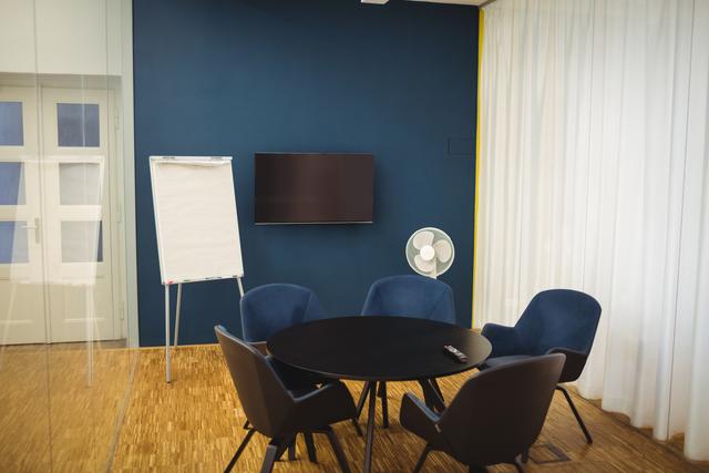 Modern empty business meeting room featuring blue chairs around a round table, a flip chart, and a TV screen on a blue wall. Ideal for illustrating professional workspaces, corporate environments, or modern office settings. Suitable for business presentations, office design concepts, and corporate training materials.
