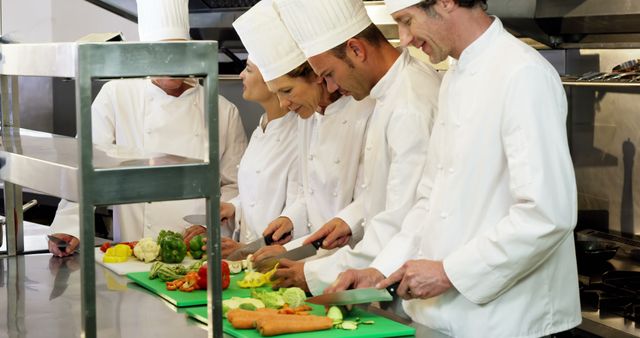 Chefs in white uniforms and hats chopping assorted vegetables on green chopping boards in a professional kitchen. Ideal for use in content related to culinary arts, teamwork in cooking, professional training, restaurant industry, and food preparation. This image showcases collaborative cooking and professional kitchen environment.