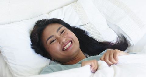 Asian woman relaxing in bed smiling while holding white duvet in soft daylight morning