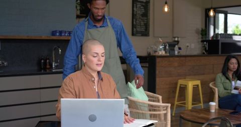 Barista approaching customer working on laptop in bright rustic café with wicker seating