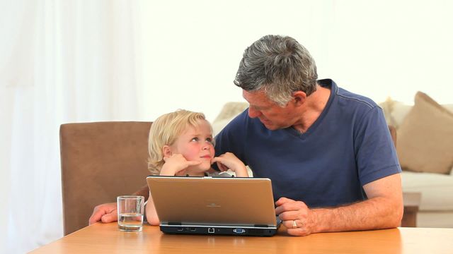 A heartwarming scene of a grandfather engaging with his grandson over a laptop at a table. The pair are sharing quality time as they explore technology together. The presence of a glass of water and cozy background enhances the feeling of a nurturing family environment. Ideal for use in advertisements promoting family computing activities, educational apps for children, or articles on intergenerational bonding and digital literacy.