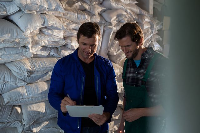 Two men are discussing work in a factory warehouse, surrounded by stacked bags. One is holding a clipboard, indicating they are reviewing or planning tasks. This image can be used for themes related to teamwork, industrial work, logistics, inventory management, and collaboration in a professional setting.