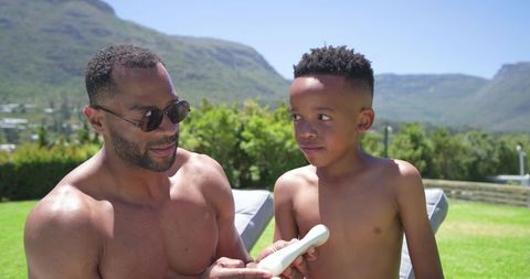 African american father applying sunscreen to son on sunny backyard lawn with mountain view