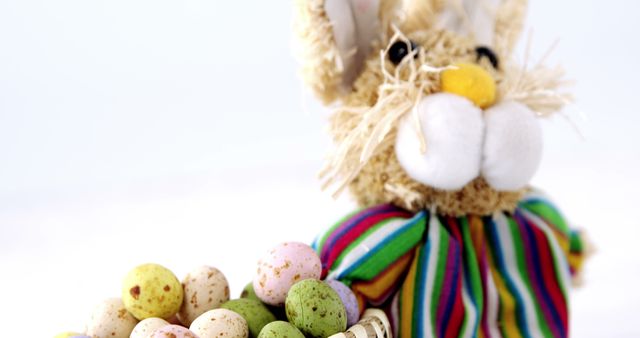 Picture depicts a cute stuffed Easter bunny in a colorful striped outfit posed with an assortment of speckled candy eggs on a white background. This image is perfect for themes involving Easter, spring celebration, holiday decorations, or children’s events. It can be used in websites, greeting cards, promotional material, or festive advertisements.