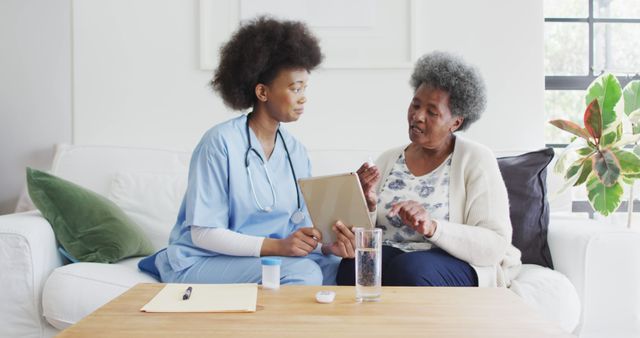 African American nurse interacting with elderly woman using digital tablet, inside home. They are sitting on a couch, highlighting a moment of care and consultation. The nurse has a stethoscope around her neck. Can be used for materials on home healthcare, geriatric care, caregiving professionals, and medical consultations.