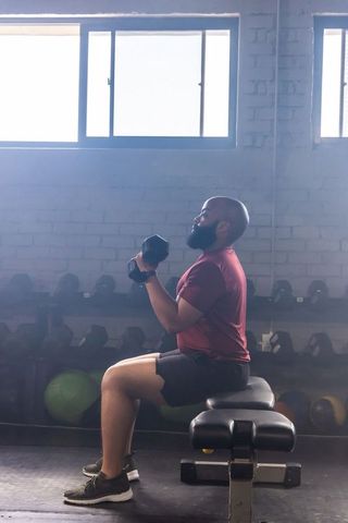 African american man sitting on bench lifting dumbbells strength training sunlit gym