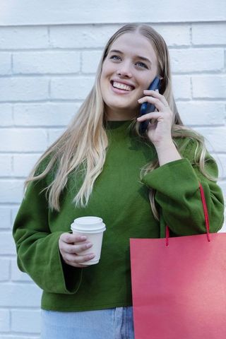 Smiling woman talking on phone holding coffee and red shopping bag by white wall