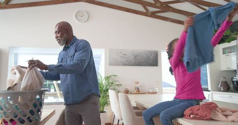Father and daughter folding laundry together in sunlit modern kitchen with wooden beams