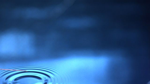 This visual capture of a syringe releasing water into a pool creates a fascinating ripple effect on a blue background, suitable for illustrating concepts related to scientific research, precision in medical procedures, or the intricate details of water dynamics. It can be used in publications and presentations about healthcare or physics of fluids.