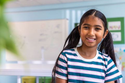 Smiling indian schoolgirl facing camera in bright classroom with whiteboard and plants