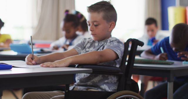 Young boy in wheelchair writing at a desk in a classroom setting. Represents concepts of inclusive education, diversity, and determination. Suitable for educational websites, blogs about disability inclusion, school brochures, and advocacy for accessible schools.