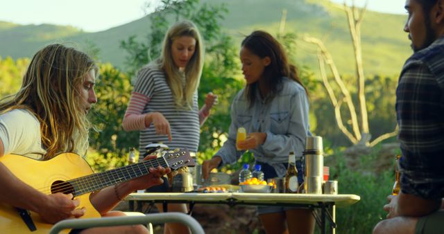 Group of young friends having an outdoor picnic in a scenic natural setting with one playing an acoustic guitar. Perfect for depicting friendship, social gatherings, leisure activities, and summer enjoyment. Useful for advertising travel, camping equipment, or lifestyle products aimed at young people.