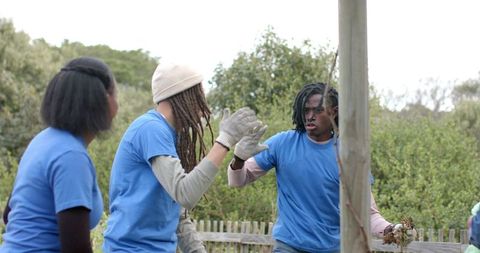 Diverse volunteer garden team high-fiving with gloves while sharing plant clippings by fence