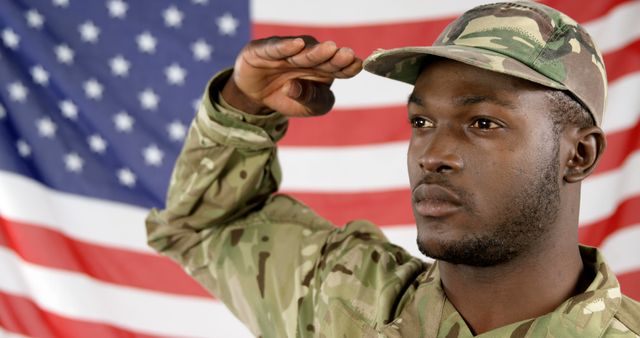 An African American soldier in military uniform salutes in front of an American flag, with copy space. His expression conveys seriousness and patriotism as he performs a gesture of respect and honor.