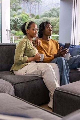 African american couple relaxing on sofa with mugs and phone in cozy sunlit living room