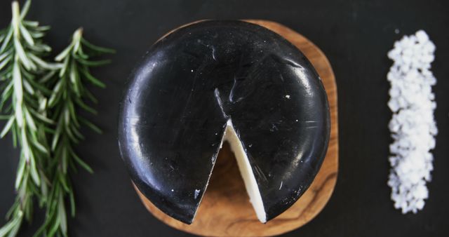 A wheel of black cheese with a piece cut out is presented on a wooden board, with copy space. Rosemary and a sprinkle of coarse salt add a culinary touch, suggesting a gourmet food setting or cheese tasting experience.