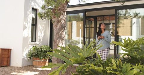 Mid-adult asian woman enjoying morning coffee on sunlit courtyard patio near glass doors