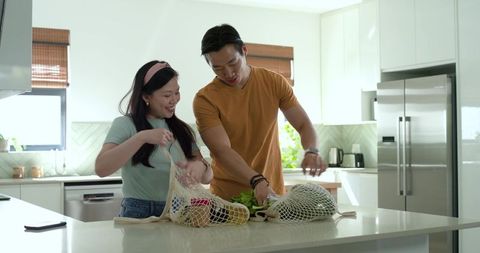 Asian couple unpacking reusable mesh grocery bags at sunlit modern kitchen island with fresh produce