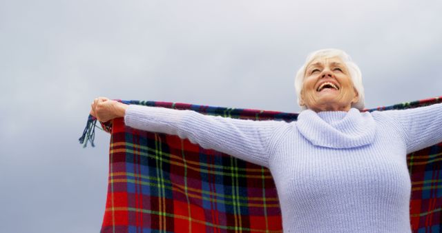 A senior Caucasian woman is holding a colorful blanket up in the air, with copy space. Her joyful expression suggests a sense of freedom or happiness, enjoying the outdoors.