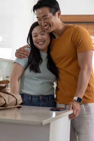 Asian couple hugging and smiling at kitchen island leaning with grocery bag and smartphone