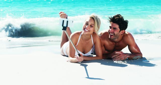 Couple lying on sandy beach near waves, using a selfie stick to take a picture. Both smiling widely and enjoying the sunny, relaxing atmosphere. Ideal for travel promotions, beach-themed advertisements, vacation brochures, and lifestyle blogs.