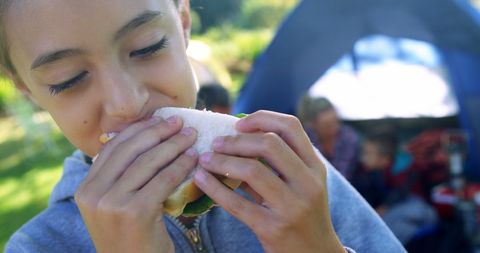 young girl savoring a sandwich outside tent. background shows family camping, representing outdoor adventure and bonding. suitable for themes of family activities, outdoor meals, and nature relaxation.