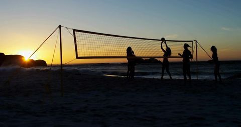 Five women playing beach volleyball at sunset silhouette along coastal shoreline waves