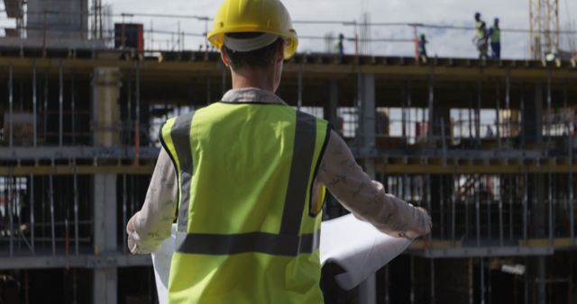 Construction worker holding and reviewing blueprints on construction site. The worker is wearing a yellow safety helmet and reflective vest, indicating engagement in active building work. The background shows a partially constructed building with metal scaffolding and additional workers in the distance. Ideal for illustrating construction industry themes, project management, engineering professions, safety protocols, and architectural design.