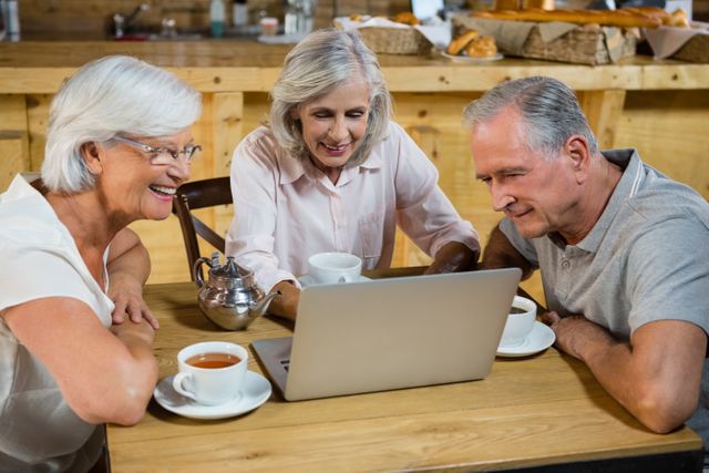 Three senior friends are gathered around a laptop in a cozy cafe, enjoying coffee and tea while engaging with technology. This image is perfect for illustrating themes of senior lifestyle, socializing, and the use of technology among older adults. It can be used in advertisements, blogs, or articles focused on senior living, technology adoption by the elderly, and social activities for retirees.