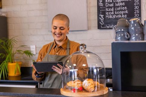 Smiling barista using tablet at cozy cafe counter with pastry dome and chalkboard menu