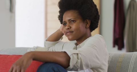 this depicts an african american woman sitting on a cozy red sofa, looking lost in thought. ideal for articles about relaxation, home living ideas, or mental health awareness. suits lifestyle blogs, home décor studies, or mindful living themes.