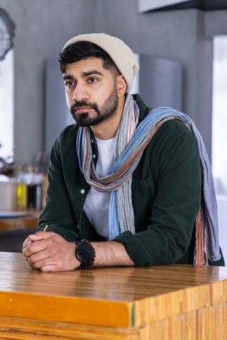 Bearded man leaning on wooden counter in modern cafe wearing beanie and multicolored scarf