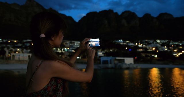 Woman standing by water at twilight, capturing scenic city and mountain view with her smartphone. Ideal for use in travel brochures, technology advertisements, social media illustrations, and tourism websites.