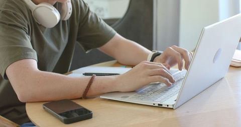 Teen male typing on slim white laptop at round wooden table with headphones and smartphone