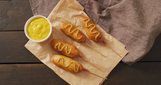 Corn dogs lying on brown paper with mustard on top, next to a bowl of mustard on a wooden table. Ideal for content about street food, snack ideas, or fast food options. Suitable for food blogs, recipe sites, or promotional material for food events.