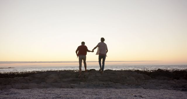 Couple is holding hands while standing on a rocky beach during sunset, expressing their love and connection. Ideal for use in travel blogs, romantic getaway advertisements, relationship coaching materials, or any promotion focusing on love and togetherness.