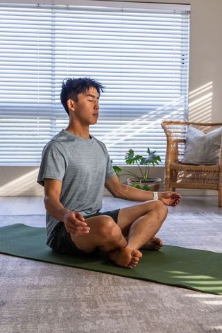 Young asian man meditating cross-legged on green yoga mat in sunlit minimalist living room