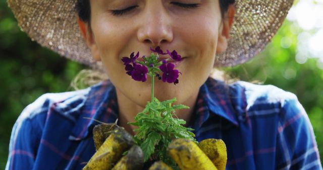 A young Caucasian woman wearing a straw hat and gardening gloves is smelling purple flowers, with copy space. Her closed eyes and content expression suggest a moment of enjoyment in nature.