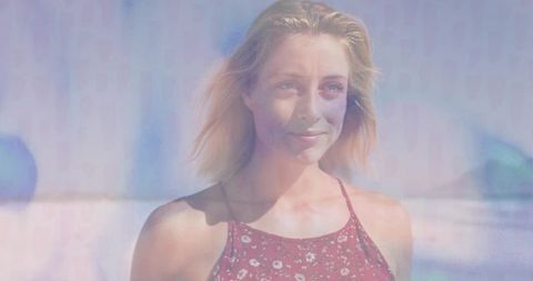 Smiling woman standing at sunlit shoreline wearing red floral dress, pastel beach portrait