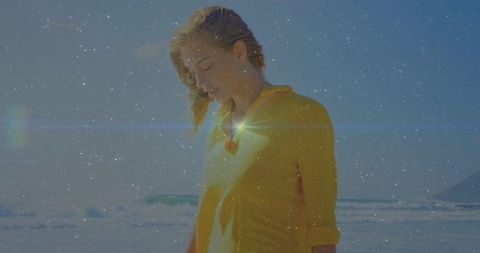 Contemplative woman standing on sunlit beach wearing yellow top with pendant and lens flare